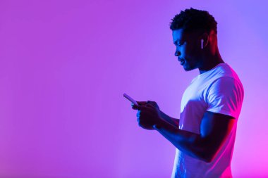 Side view of young black man in earphones listening to music, using new mobile app on smartphone in neon light, free space. Cheerful African American guy enjoying new playlist or audio book