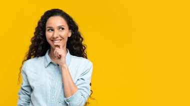 Happy thoughtful young woman in casual outfit looking at empty space over yellow studio background, banner design. Positive millennial female offering free place for ad or promo