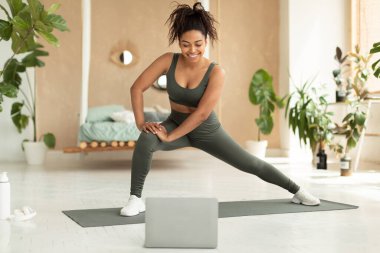 Good looking african american lady enjoying online fitness class while staying home, looking at laptop screen, doing workout in bedroom interior. Sports lifestyle concept