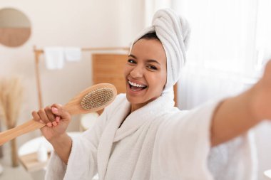 Cheerful Woman Having Fun Caring For Body Singing Holding Wooden Massage Brush Like Microphone Standing In Modern Bathroom Indoor, Posing With Towel On Head. Beauty And Bodycare Cosmetics