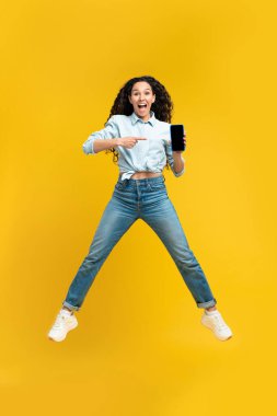 Excited millennial lady pointing at smartphone with empty screen, jumping up in air, offering space for mobile app or website on yellow studio background, mockup. People and gadgets concept