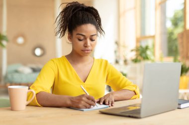 Focused black female student taking notes while watching online lecture on laptop, learning online at home, sitting at desk. African american freelancer writing article using pc computer