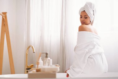 Beauty And Spa. Attractive Female With Wrapped Towel On Head Posing Sitting Back To Camera In Modern Bathroom Indoor. Bodycare And Skincare Cosmetics Concept