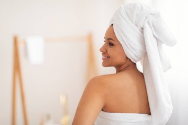 Wellness And Spa. Young Lady With Wrapped White Towel On Head Posing Standing Back To Camera Looking Aside In Modern Bathroom At Home. Beauty Routine Concept