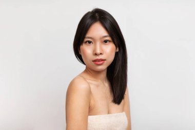 Pretty young korean lady standing wrapped in towel over white background. Portrait of asian woman with smooth skin and dark hair posing after bath