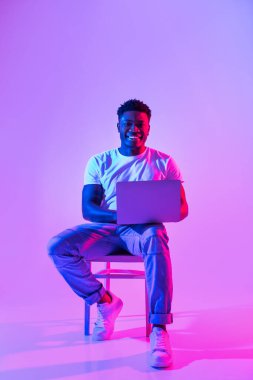 Full length of happy millennial black guy with laptop pc sitting on chair, studying or working online in neon light, copy space. Cheery African American man having remote business meeting on web