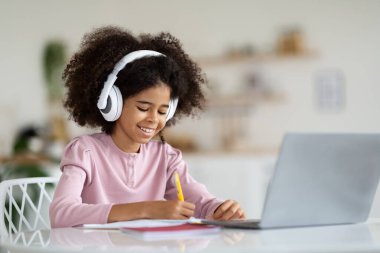 Cheerful african american schooler cute preteen girl with bushy hair sitting at table in front of laptop, using wireless headset, taking notes, doing homework at home, copy space