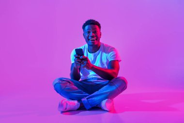 Full length of excited young African American guy sitting cross legged with cellphone, posting in social media, chatting on web in neon light, copy space. Millennial black man using smartphone