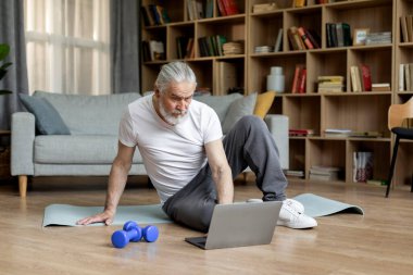 Muscular elderly man in sportswear sitting on fitness mat in living room, choosing online workout class, using laptop and barbells, copy space, full length. Fitness studio on Internet for seniors
