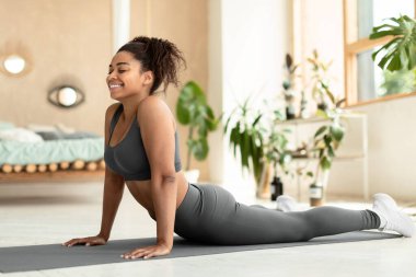Working out at home. Fit black lady stretching, doing Cobra exercise or standing in upward-facing dog posture on fitness mat, wearing sportswear, copy space