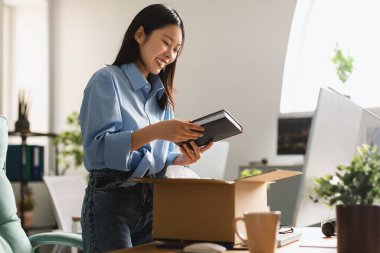 Cheerful Asian Businesswoman Unpacking Box Holding Book Standing In Modern Office Indoors. Business Literature, Shopping And Delivery Service Concept. Side View