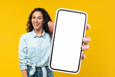 Excited Middle Eastern Woman Showing Large Cellphone With Blank Screen Recommending Mobile Application Standing Over Yellow Studio Background, Smiling To Camera. Mockup