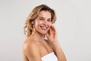 Self-Care Concept. Closeup Portrait Of Joyful Middle Aged Female With Beautiful Skin Standing Wrapped In Bath Towel Over Light Grey Background In Studio, Attractive Lady Smiling At Camera, Copy Space