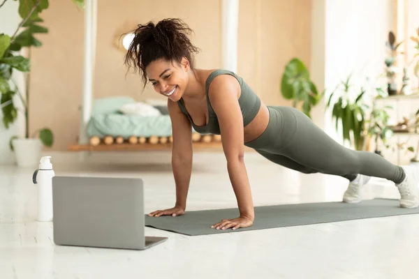 Excited athletic african american lady exercising at home, sporty black woman enjoying online yoga class, using laptop computer. Healthy lifestyle concept
