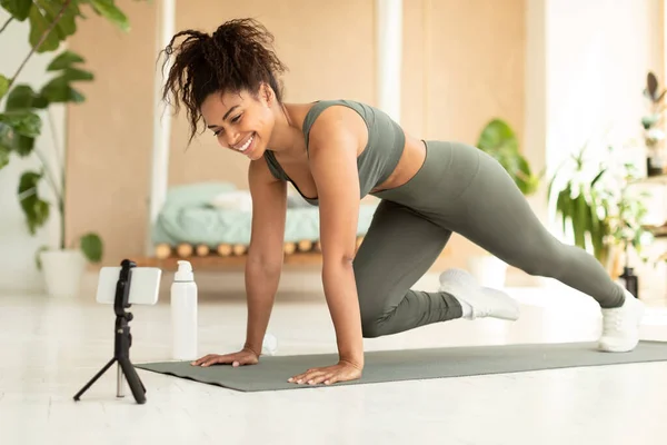 Online workout. Fit african american female doing cross body mountain climbers exercise on yoga mat, looking at smartphone on tripod, recording video for blog or having chat with coach