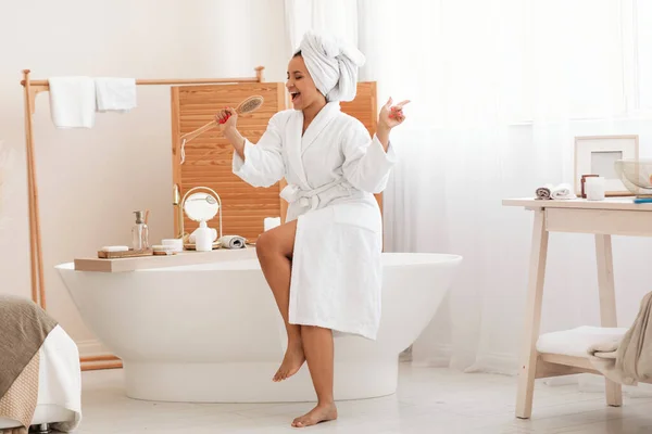 Happy Female Singing Holding Wooden Brush Caring For Herself And Having Fun Posing With Wrapped Towel On Head And Wearing White Bathrobe Sitting In Modern Bathroom Indoor