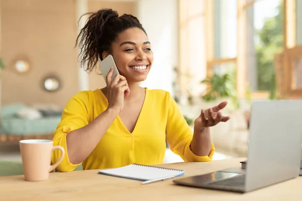 Portrait of excited black woman working from home, talking on smartphone and using laptop, sitting at desk at home. Happy lady having phone conversation, making corporate call