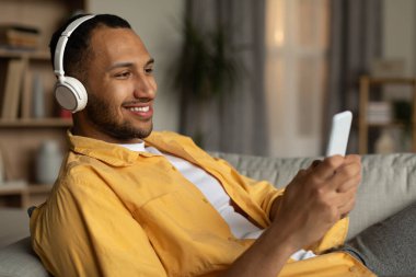 Joyful young black guy in headphones lying on couch and listening to music online on cellphone at home. Cool millennial African American man watching video on smartphone, using wireless headset