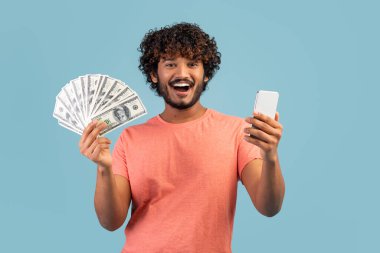 Happy emotional wealthy young eastern guy in casual holding cell phone and bunch of cash dollars, posing over blue studio background, trading online on stocks and markets, won lottery, mockup