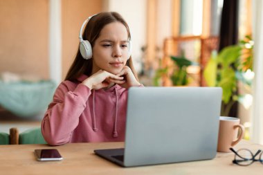 E-education, online lessons concept. Focused teen girl in headset looking at laptop screen, watching online lesson or listening to tutor, sitting at table at home, free space