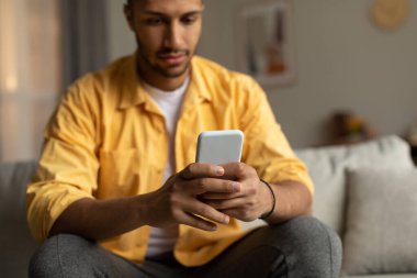 Serious young African American guy sitting on couch with smartphone, chatting online, browsing social network, watching video, messaging with friend at home, selective focus. Copy space