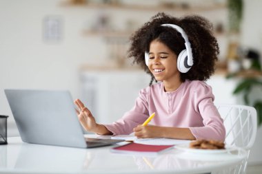Positive black kid school girl with bushy hair having online party with friends while staying home, sitting at desk, drawing, using wireless headset, waving and smiling at computer screen, copy space