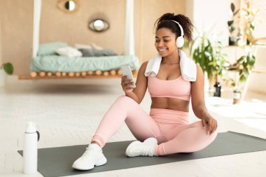 Positive fit black woman with towel on shoulders and wireless headphones sitting on fitness mat and listening to music while resting during home workout, free copy space