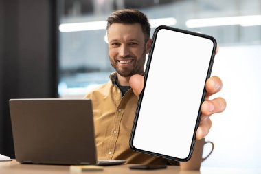 Business Offer. Smiling Millennial Man Demonstrating Big Blank Smartphone While Sitting At Desk With Laptop In Office, Cheerful Young Businessman Recommending Mobile App Or Website, Collage, Mockup