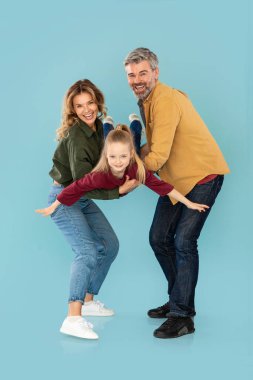 Cheerful Parents Playing With Little Daughter Holding Her In Arms Posing On Blue Background. Studio Shot Of Family Having Fun Together, Kid Spreading Hands Like Plane. Vertical