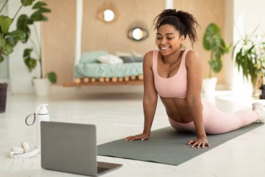 Happy african american woman stretching back and watching online tutorials on laptop, excercising in bedroom on yoga mat, standing in cobra or upward-facing dog pose