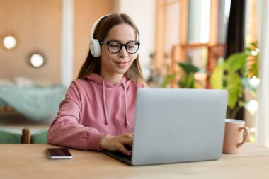 Pretty teen girl in wireless headset using laptop, interested happy student typing on keyboard looking at pc screen, having online course or doing homework