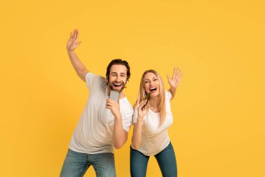 Satisfied glad millennial caucasian family in white t-shirts in headphones sing at smartphones, isolated on yellow background. Superstars and imaginary microphone, fun together and audio app, studio