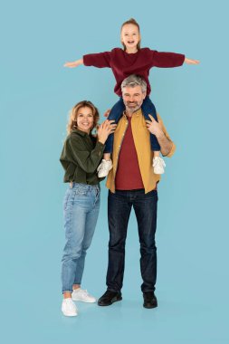 Mother And Father Posing, Holding Little Daughter On Shoulders Spreading Arms Smiling To Camera On Blue Studio Background. Happy Family Concept. Vertical Shot