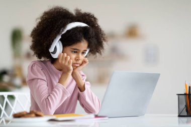 Exhausted african american school girl suffering while doing homework, tired from studying at home, getting ready before exams, stressed with difficult task, using laptop and wireless headphones