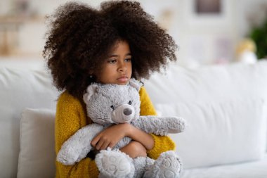 Upset preteen african american girl with curly bushy hair sitting on couch alone, embracing teddy bear, looking at copy space. Kid feeling lonely at home, missing parents or friends