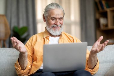Positive good-looking old man sitting on couch in cozy living room, using modern laptop at home, having video chat, looking at computer screen and gesturing, copy space. Telecommunication concept