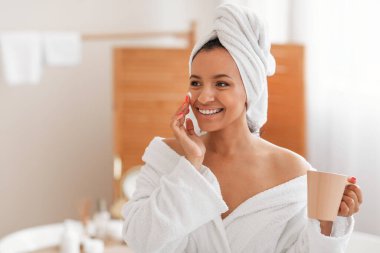 Facial Skincare. Happy Young Female Applying Moisturizer On Face Moisturizing Skin Holding Coffee Cup Standing Smiling Looking Aside In Modern Bathroom Indoors. Beauty And Cosmetics