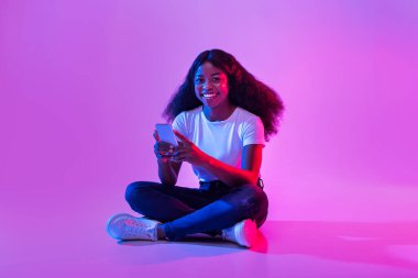 Full length of young African American female sitting cross legged with mobie phone, surfing internet, chatting online in neon light. Cool black woman using smartphone, checking email