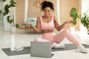 Remote trainings concept. Happy black female coach having online fitness class with laptop, talking and smiling at computer webcam while exercising on yoga mat