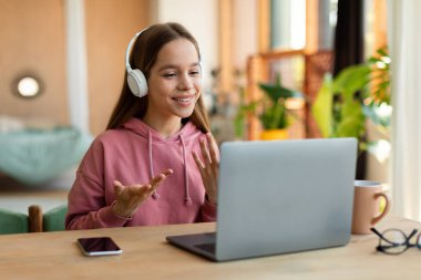 Positive schoolgirl having video call with tutor, talking to laptop webcamera, sitting at desk at home, wearing wireless headphones and gesturing. Learning and communicating online