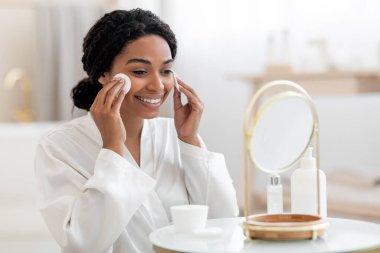 Beautiful Black Female Looking At Mirror And Cleansing Skin With Cotton Pads, Attractive Smiling African American Woman Making Daily Beauty Routine At Home, Enjoying Skincare Treatments, Copy Space