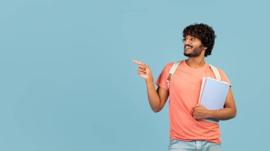 Happy young hindu guy going to college, carrying books, backpack, pointing at copy space and smiling over blue studio background, panorama. University, school, education concept