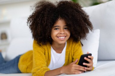 Relaxed smiling african american little girl with bushy hair lying on couch with smartphone, happy kid chatting with friends, using nice mobile application, home interior, copy space
