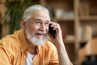 Emotional handsome long-haired bearded old man in yellow shirt having phone conversation with lover or friend, looking at copy space and smiling, sharing good news, home interior