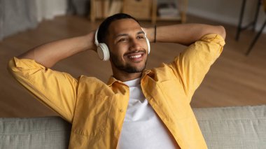 Handsome young black guy in headphones relaxing on couch, listening to music at home, banner design. Millennial African American man enjoying audio book in headset, resting indoors