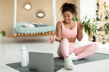Online exercise session marathon. Happy black woman sitting on floor mat and talking with coach, using pc for virtual workout, exercising at home, free space