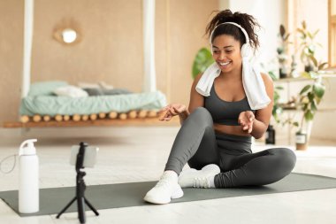 Fitness blogging concept. Fit black lady shooting online trainings using smartphone on tripod, recording video content for her sport blog, sitting on yoga mat, free space