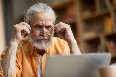 Concentrated grey-haired bearded old man in casual outfit using modern notebook, reading article, surfing on Internet, touching his eyeglasses, home interior, closeup, copy space. Eyesight, vision