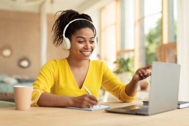 Online education concept. Happy black female student in headphones having video call via laptop, talking with tutor and taking notes, sitting at desk at home, free space
