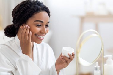 Skincare Concept. Beautiful African American Female Looking At Mirror And Applying Moisturising Face Cream, Happy Black Woman Wearing White Silk Robe Making Beauty Treatments At Home, Closeup